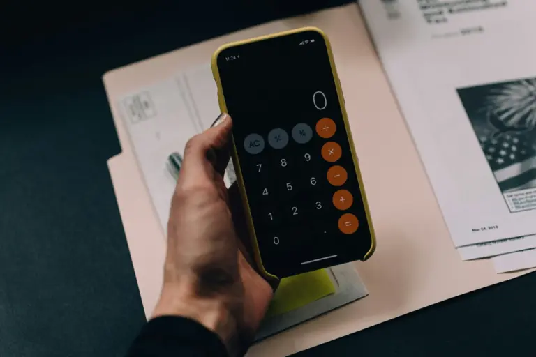 Main tenant un smartphone affichant une application de calculatrice, sur un bureau avec des documents administratifs.