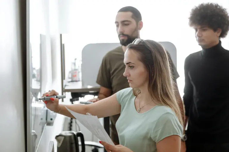 Femme écrivant sur un tableau blanc, deux hommes regardant.